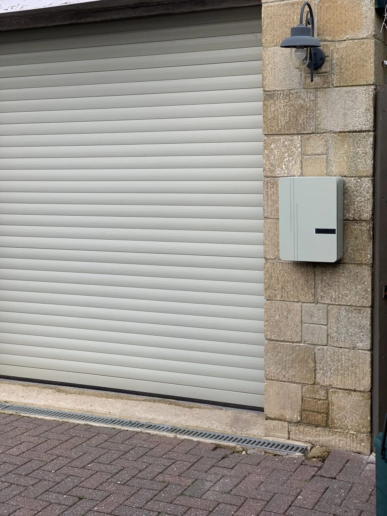 Pale grey Andersen A3 EV charger on a stone wall beside a roller garage door, below an outside lamp