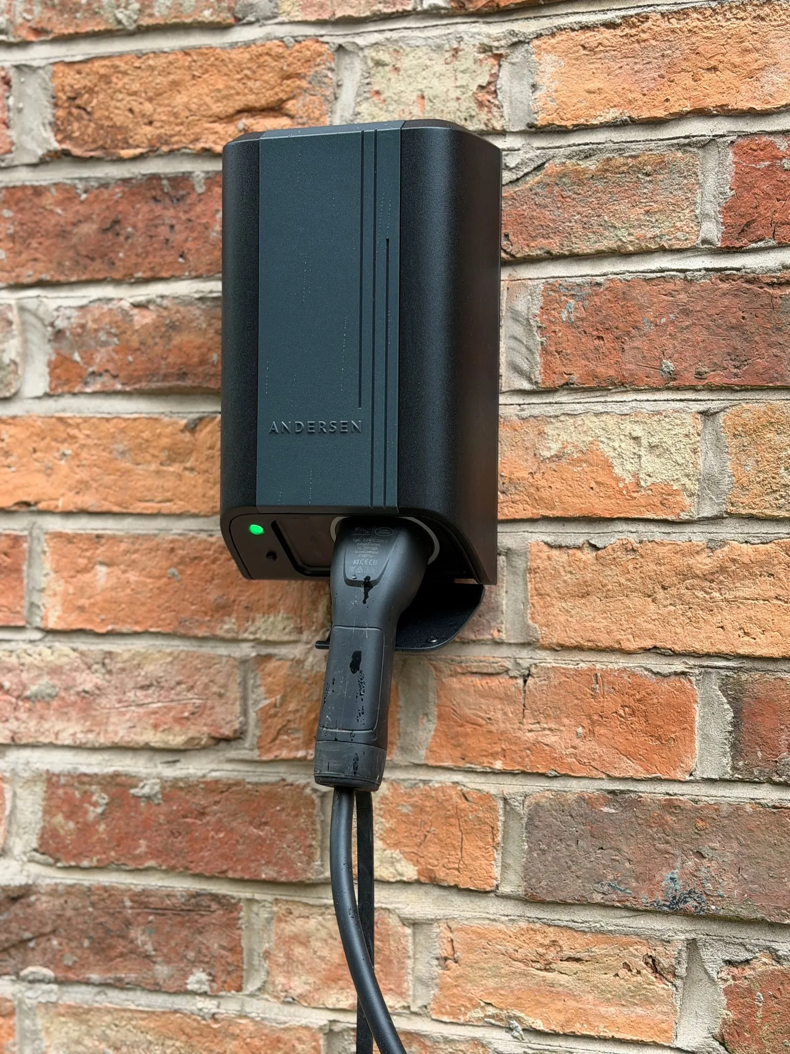 Black Andersen Quartz EV charger with a tethered connector, on a red brick wall