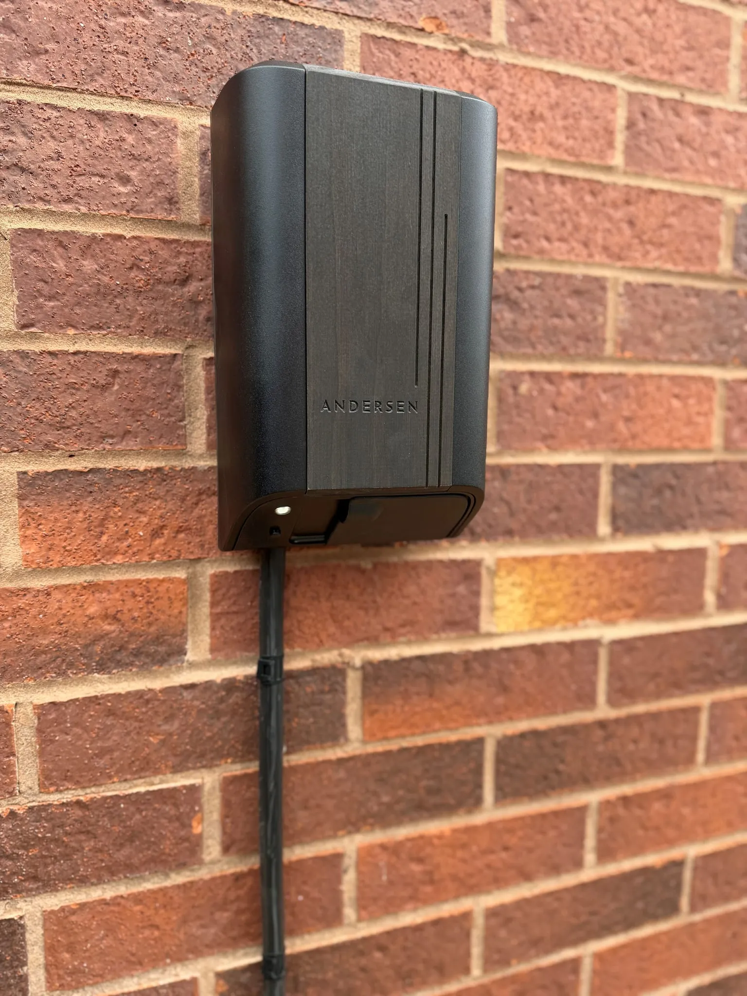 Andersen Quartz EV charger with a dark wood-effect front panel, on a red brick wall