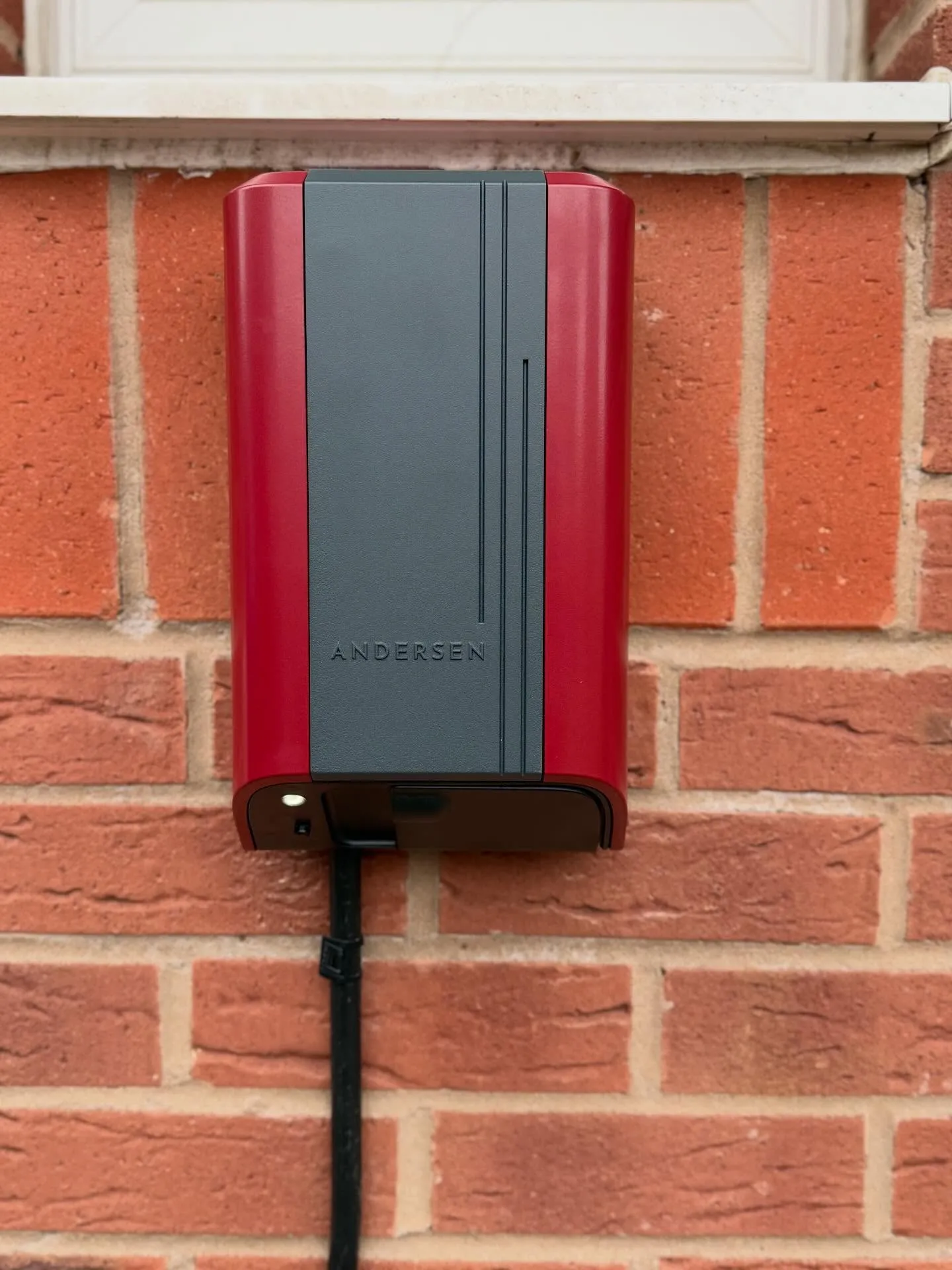 Red and grey Andersen EV charger mounted on a red brick wall below a white window sill