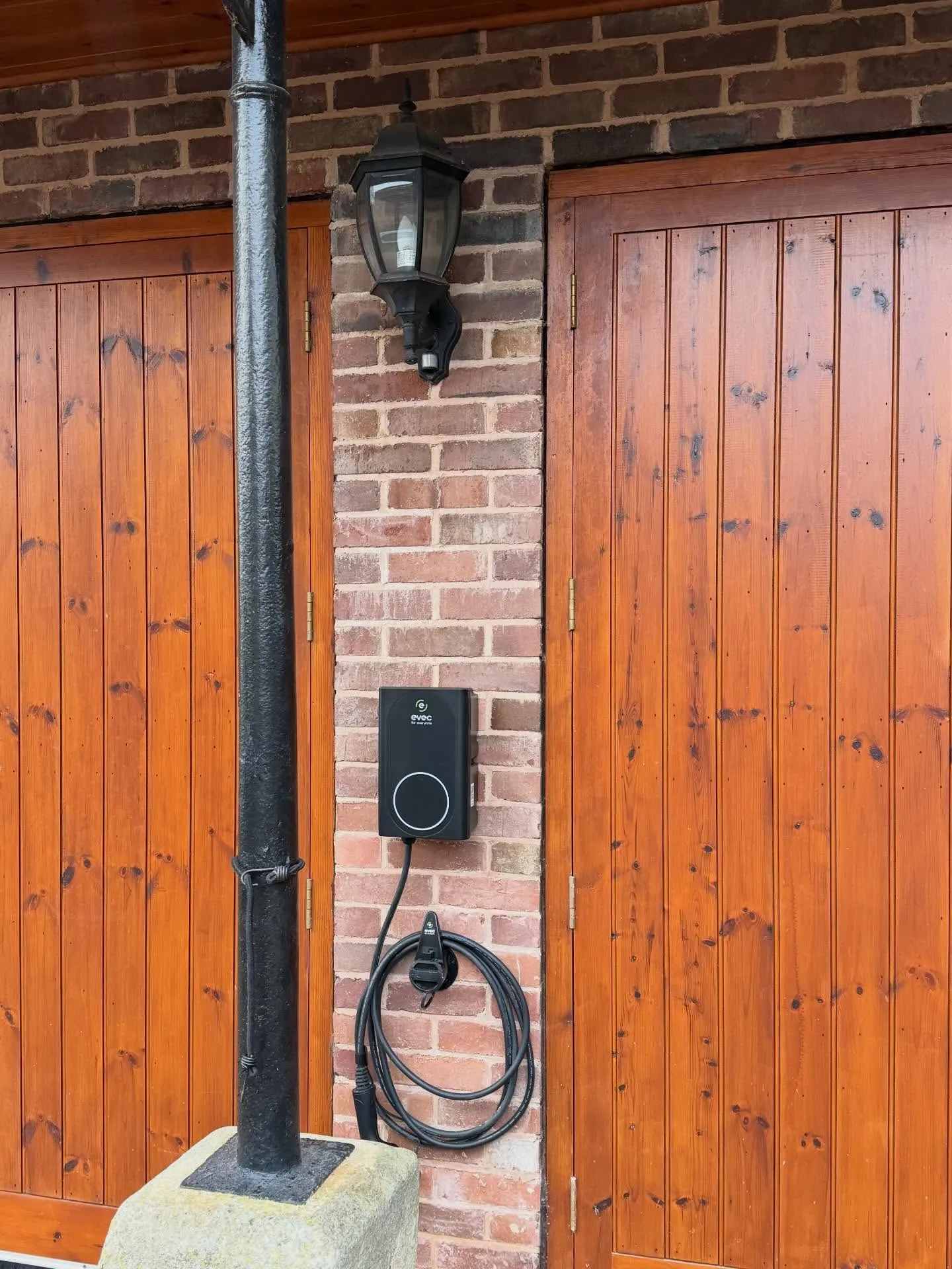 Small black Evec EV charger with a coiled cable, mounted on brickwork between two wooden garage doors below an outside light