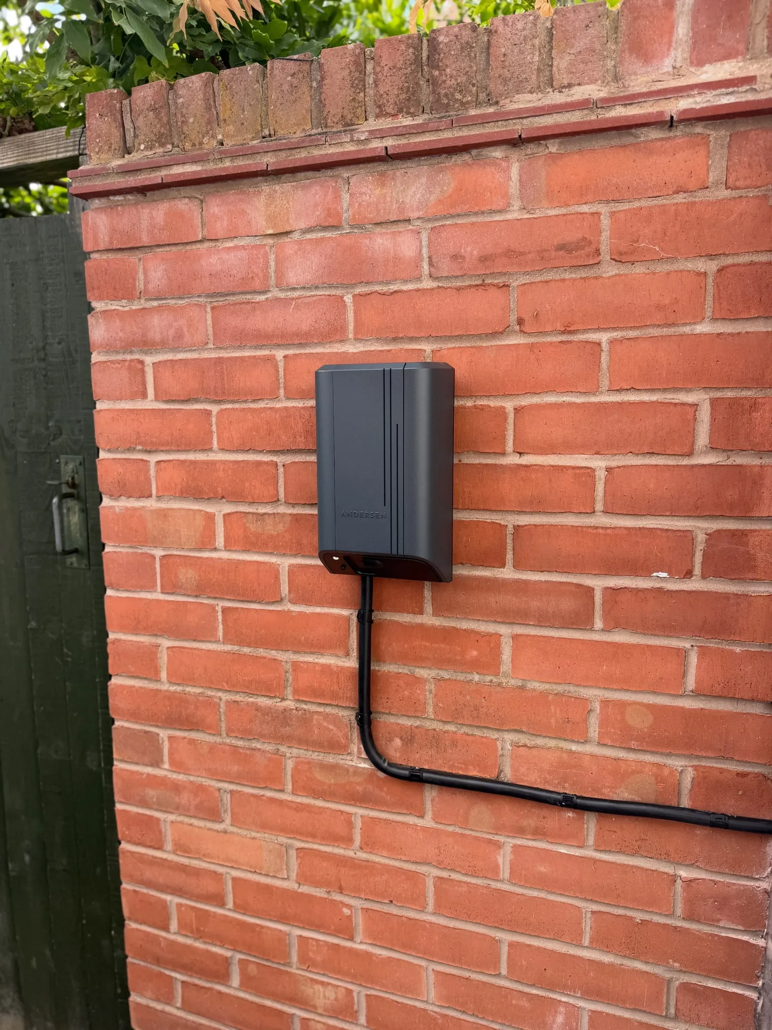 Dark grey Andersen Quartz EV charger on a brick garden wall, with its supply cable neatly clipped along the brickwork