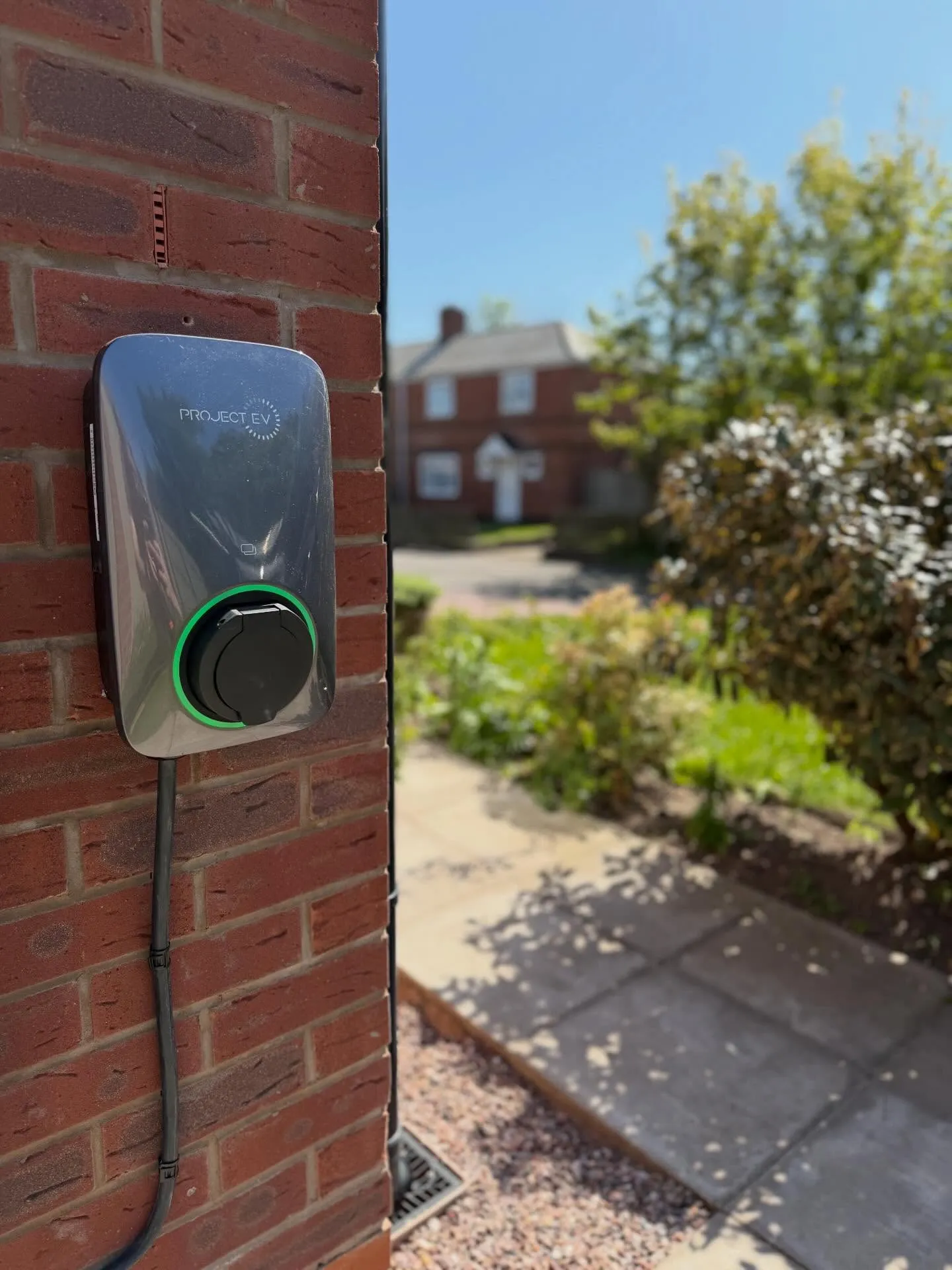 Silver Project EV charger with a green illuminated ring, mounted on a brick wall beside a garden path