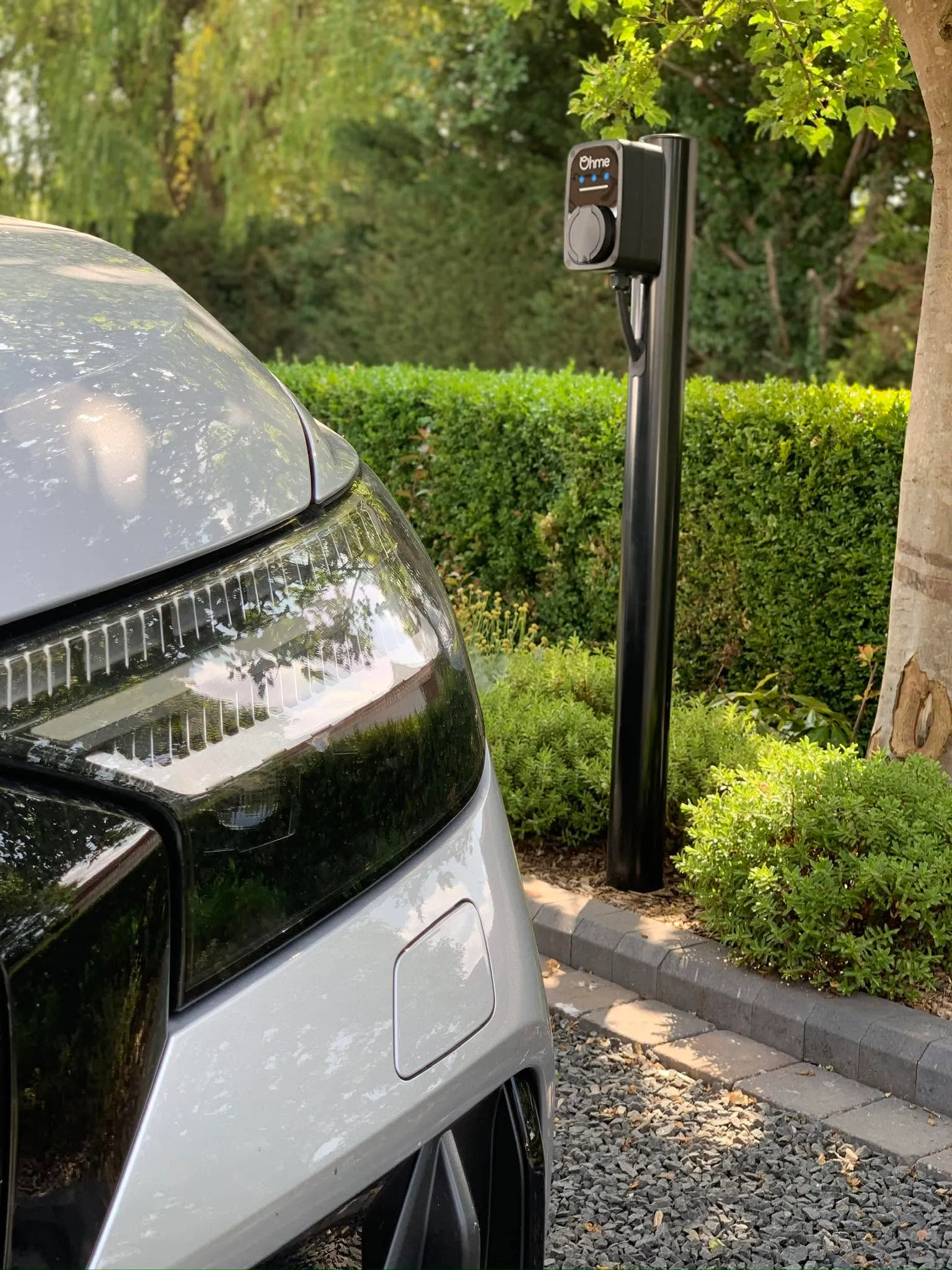 Ohme EV charger mounted on a black post beside a hedge and gravel driveway, with the front of a car in the foreground