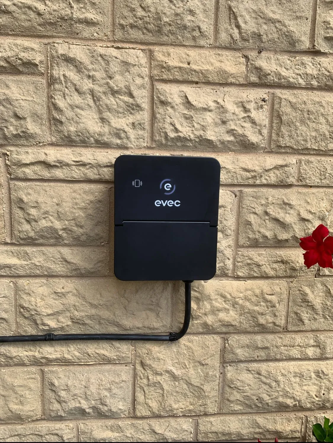 Black Evec EV charger on a buff stone wall, with its supply cable clipped along the stonework