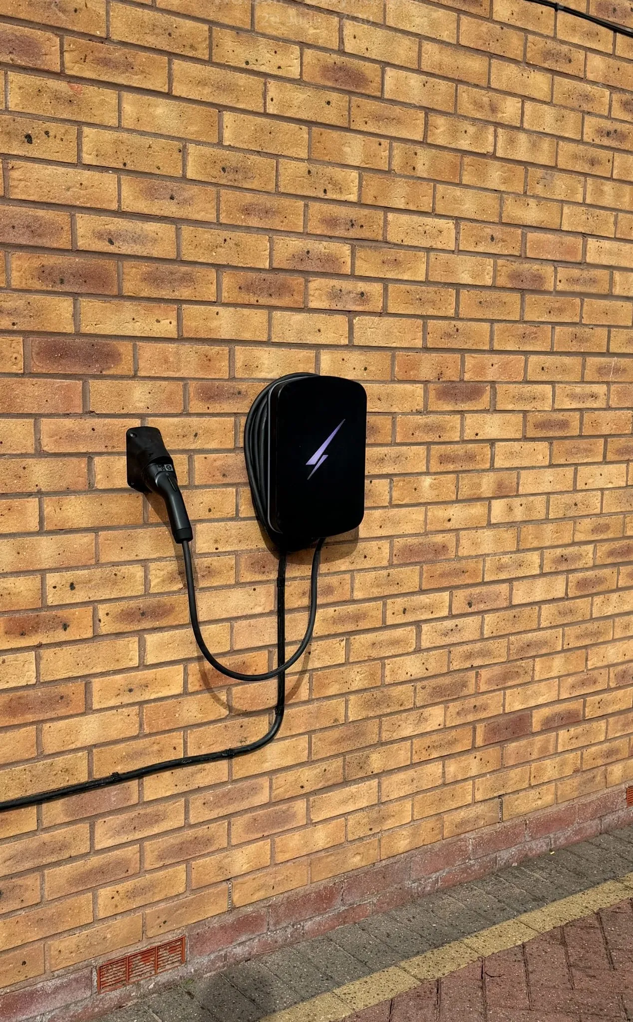 Black Hypervolt EV charger with a coiled cable and a wall-mounted connector holster, on a yellow brick wall