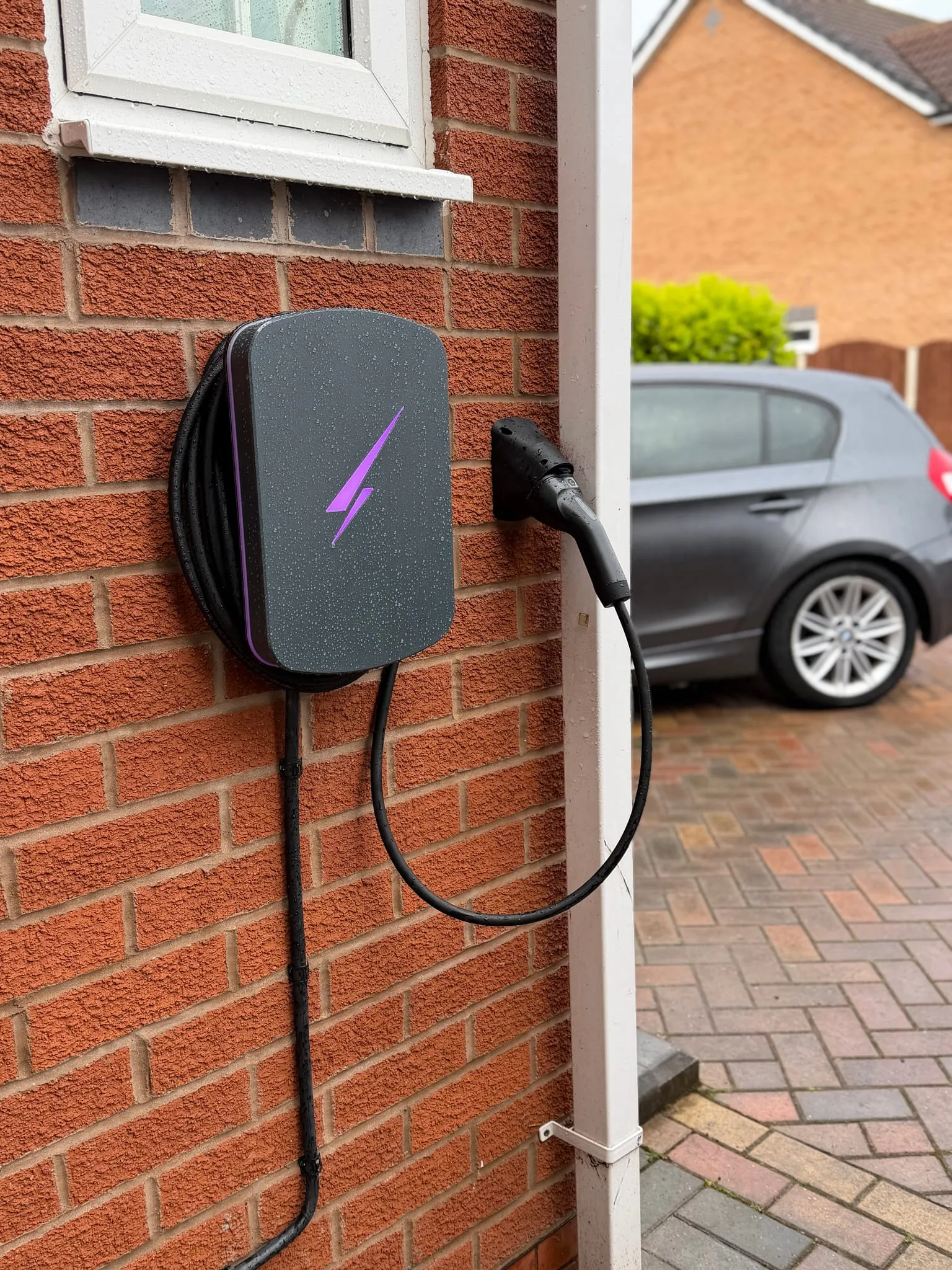 Grey Hypervolt EV charger with a purple illuminated logo on a brick wall beside a white downpipe, with a car on the block-paved driveway