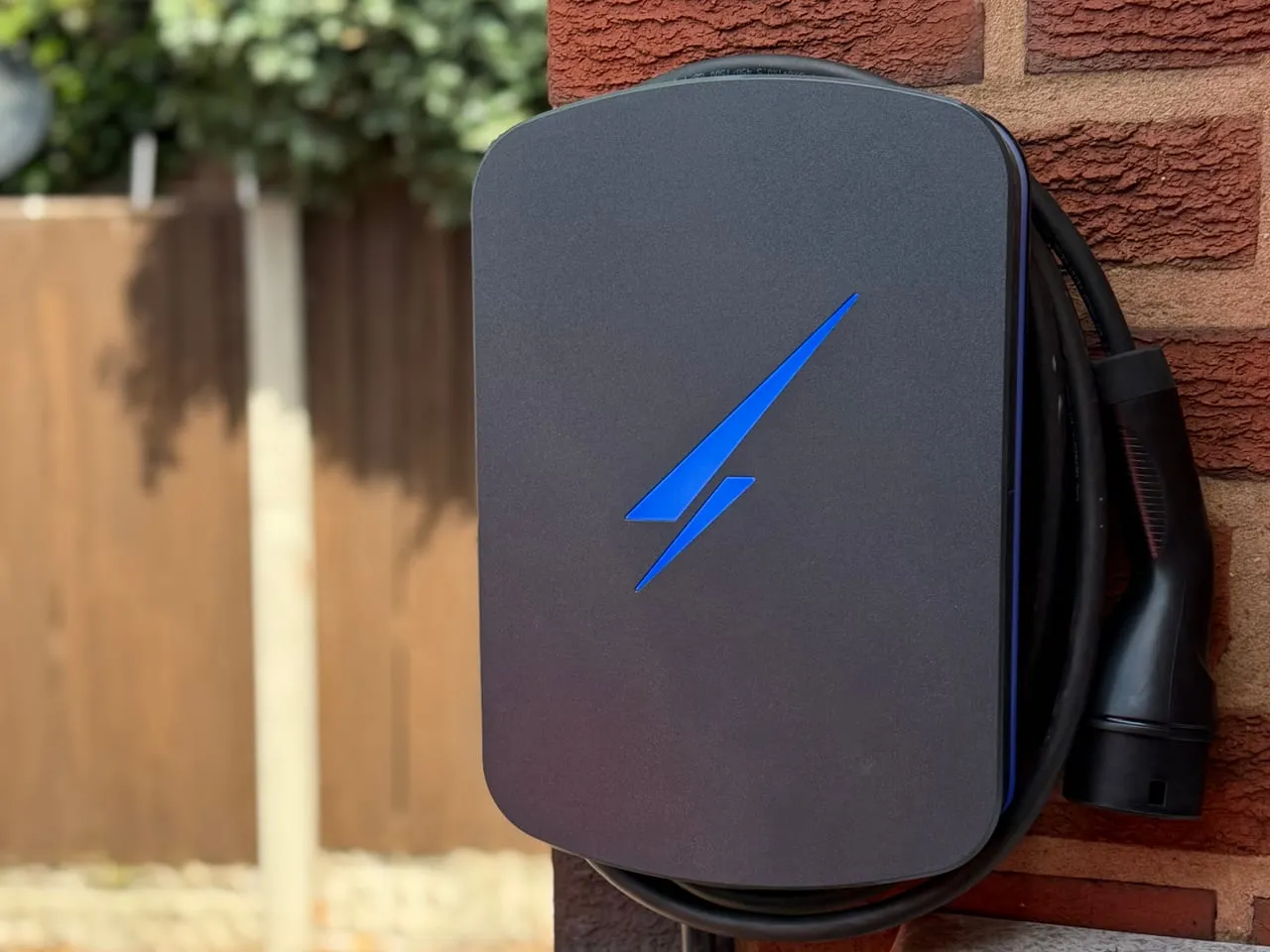 Grey Hypervolt Home 3 Pro EV charger with a blue illuminated logo and coiled cable, on a brick wall