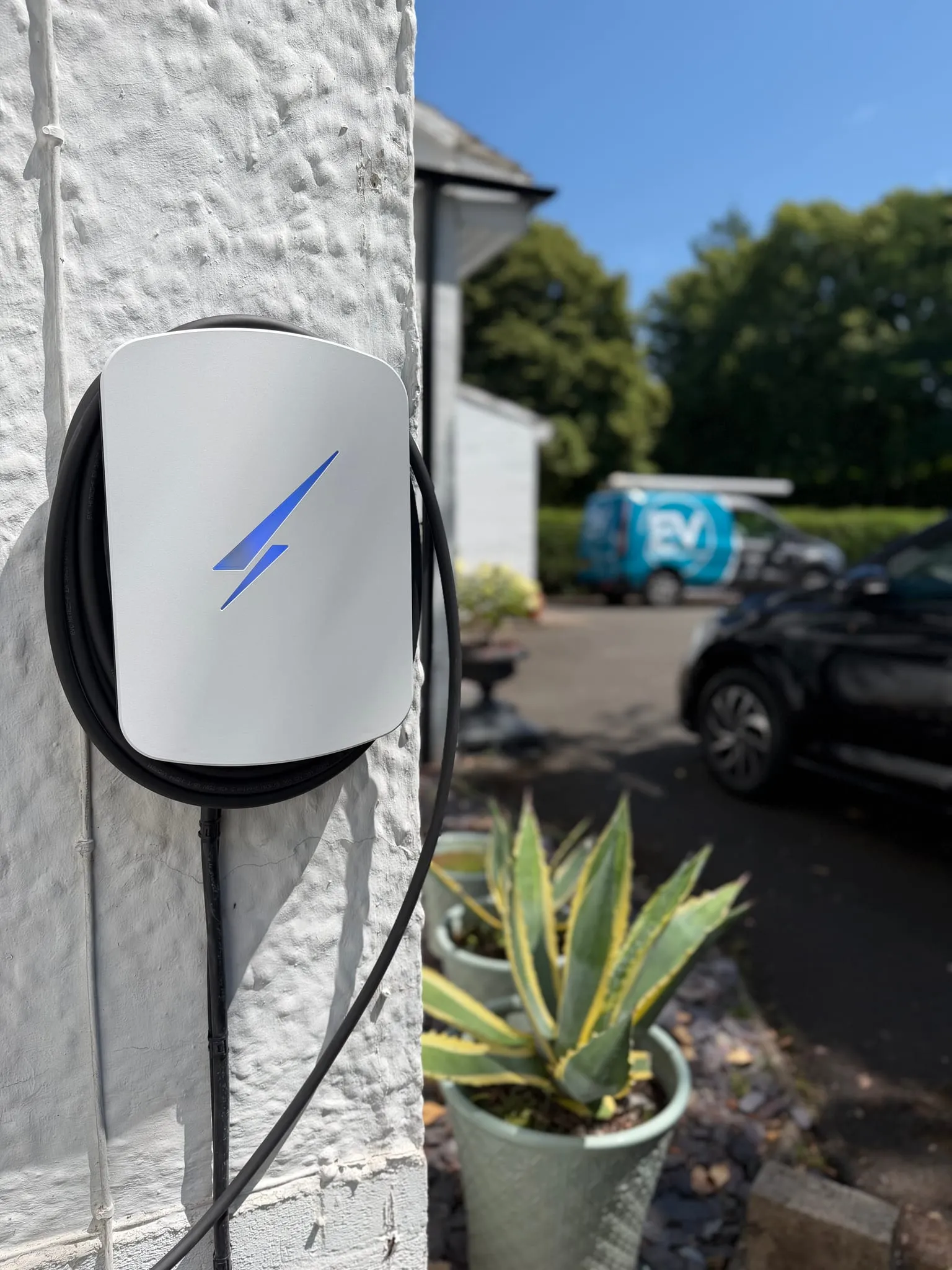 White Hypervolt EV charger with a blue illuminated logo and coiled cable on a white rendered wall, with the EV Charge Solutions van parked on the driveway behind