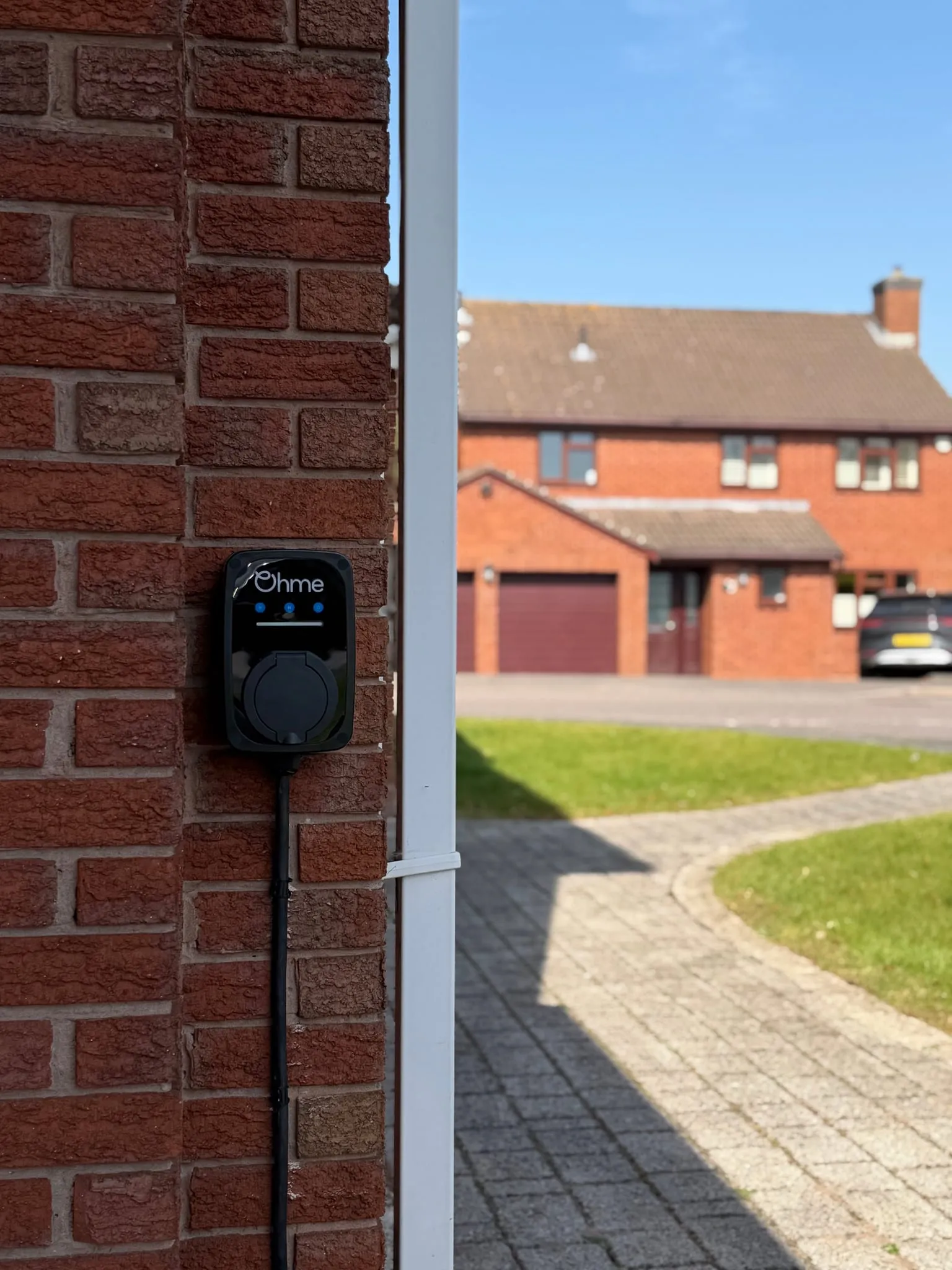 Ohme ePod EV charger on a brick pillar beside a white downpipe, looking out over a block-paved path and lawn