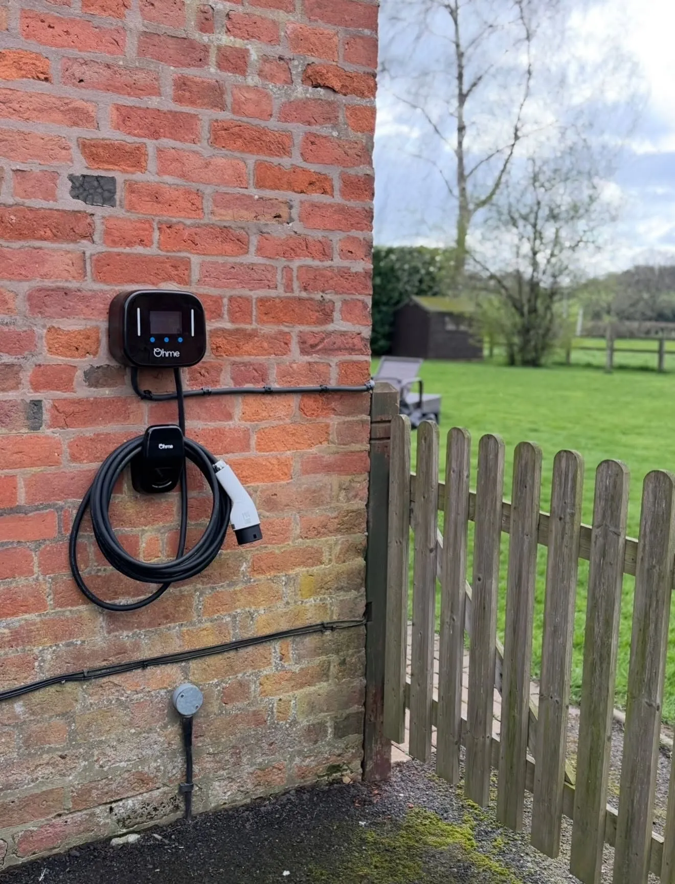Ohme EV charger with a coiled cable on a brick wall beside a wooden fence, with the supply cable neatly clipped along the brickwork