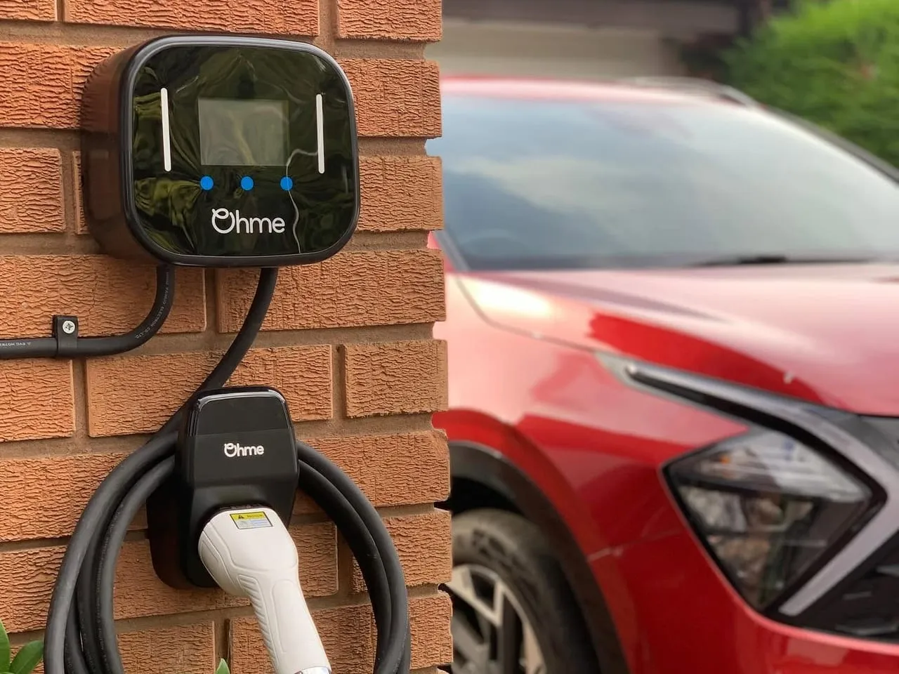 Ohme Home Pro EV charger with its cable coiled on a holder, on a brick wall beside a red car