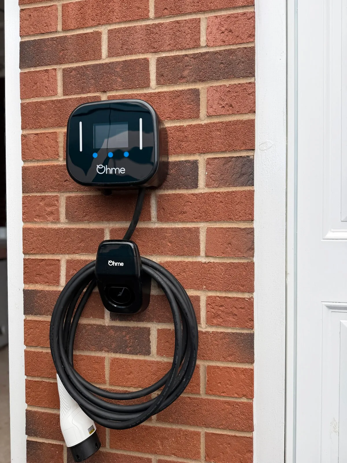 Black Ohme Home Pro EV charger with coiled cable on a brick wall beside a white door frame