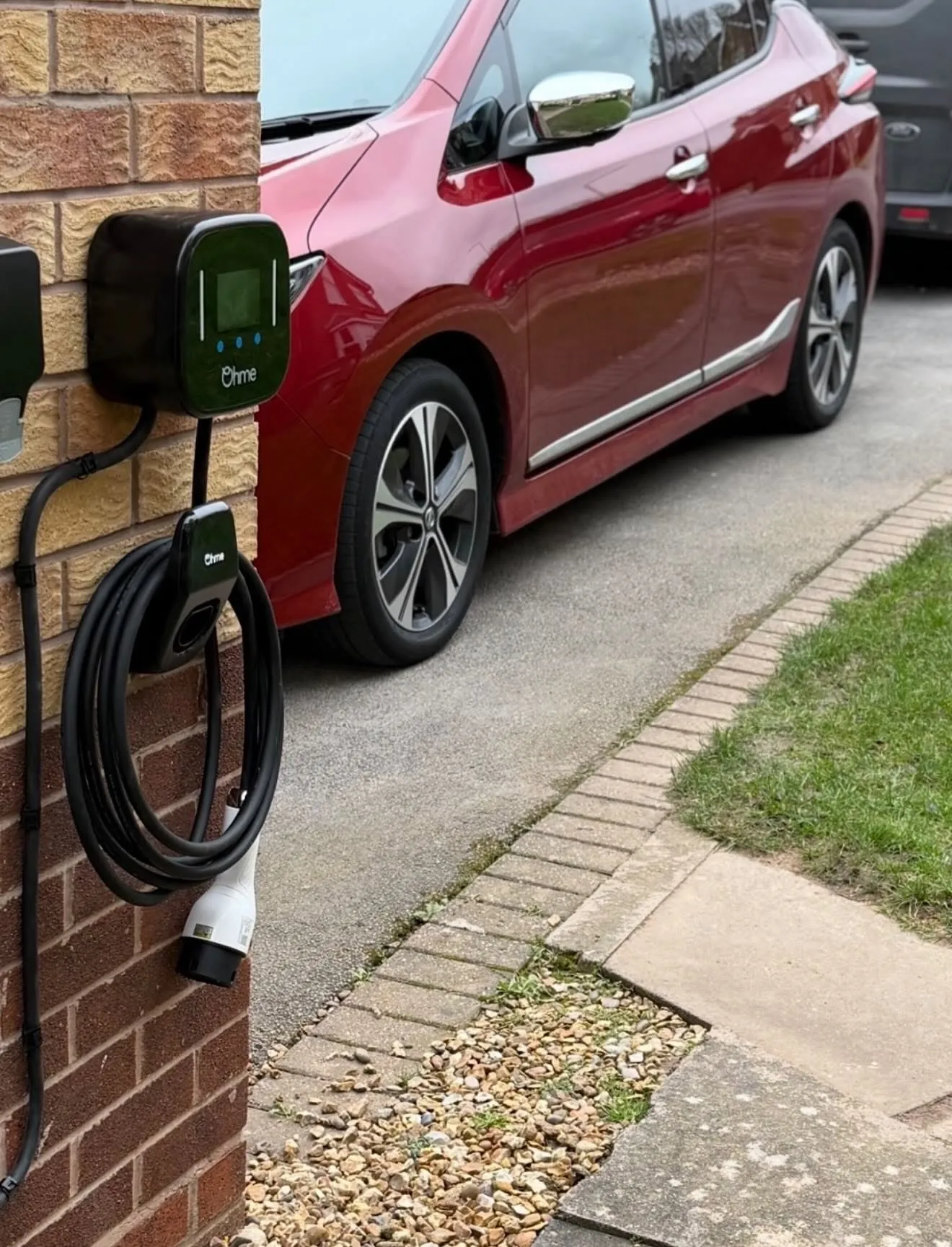 Ohme EV charger with a coiled tethered cable on a brick wall beside a paved path and driveway with a red car