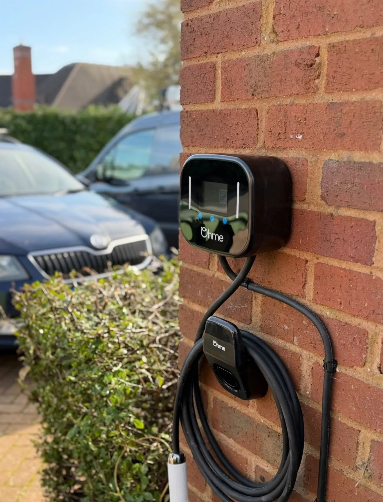Ohme EV charger with a coiled tethered cable on a brick wall beside a hedge, with cars parked on the driveway