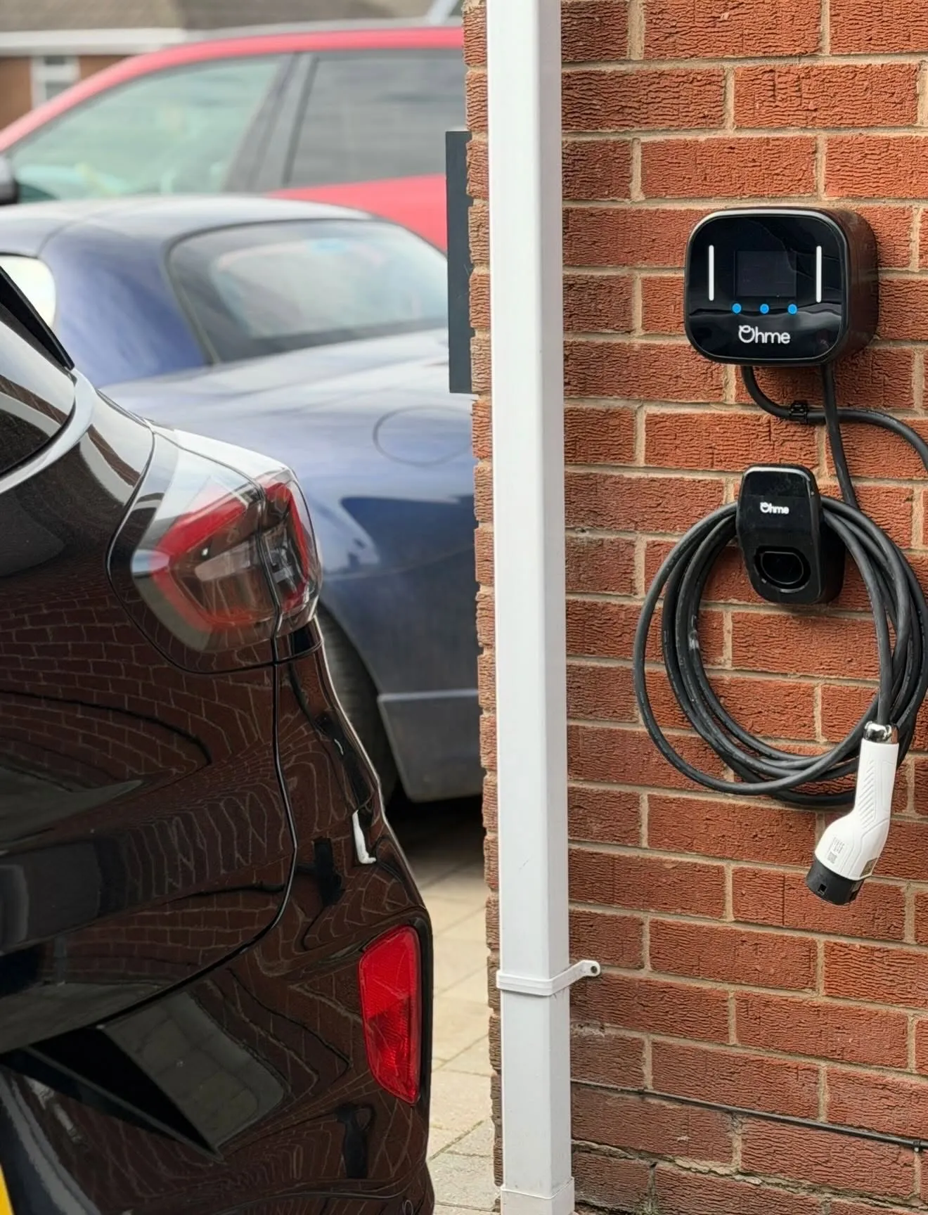Ohme EV charger with a coiled tethered cable on a brick wall beside a white downpipe, with cars parked on the driveway