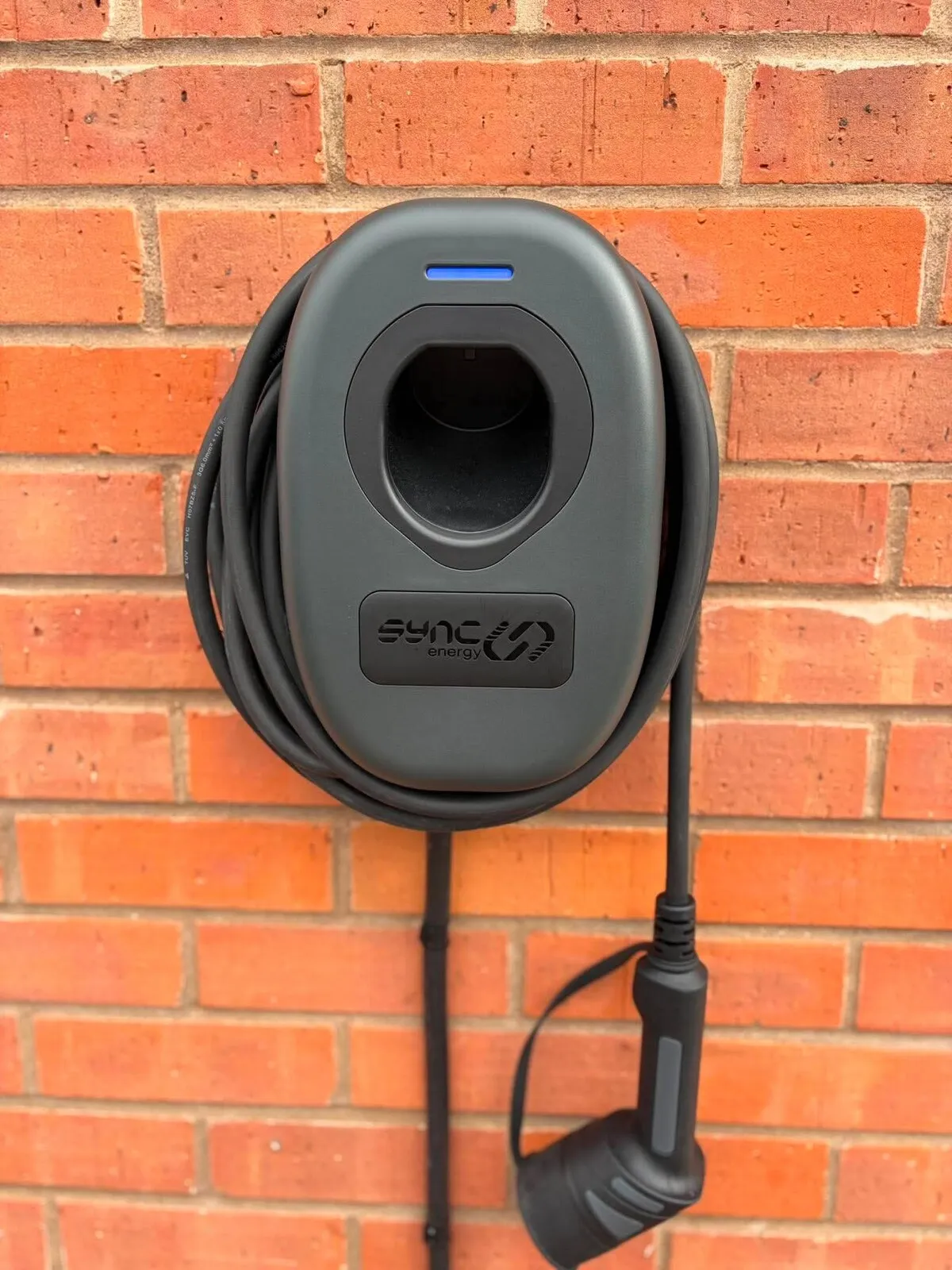 Dark grey Sync Energy Wall Charger 2 with a blue status light and coiled cable, on a brick wall