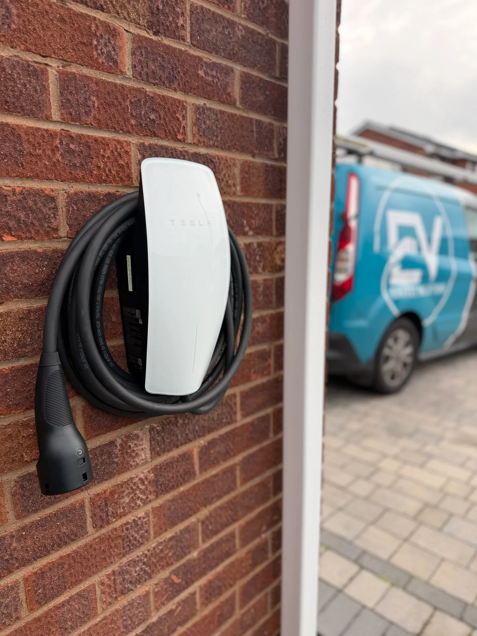 White Tesla Wall Connector with a coiled cable on a brick wall, with the EV Charge Solutions van on the driveway behind