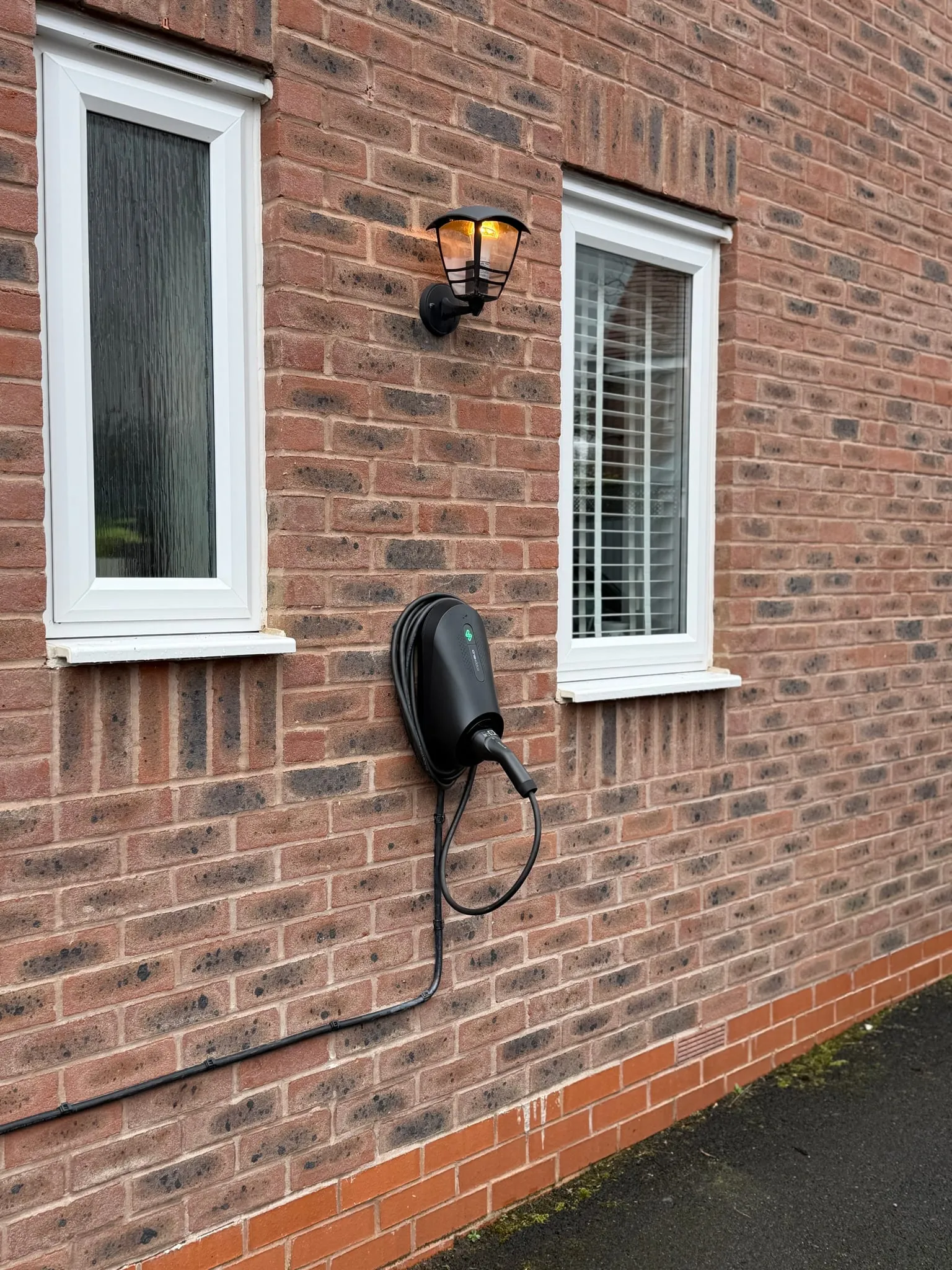 zappi GLO EV charger with a tethered cable on a brick house wall between two windows, below an outside lamp